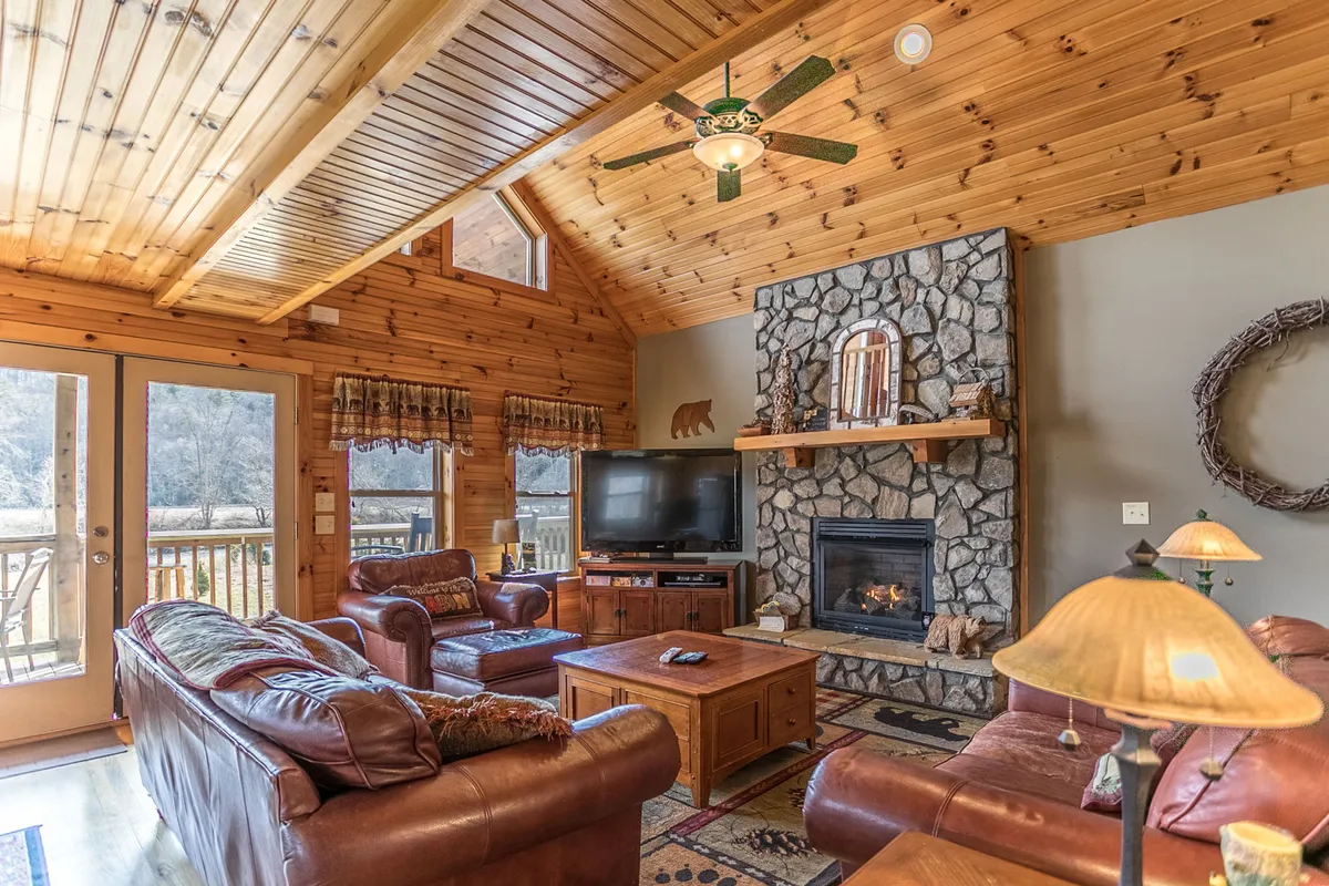 A cozy, warmly lit great room with a striking stone fireplace, wooden ceiling, leather sofas, and a coffee table. The space is perfect for gathering in winter, whether to enjoy the fire or gaze out to the deck during summer.