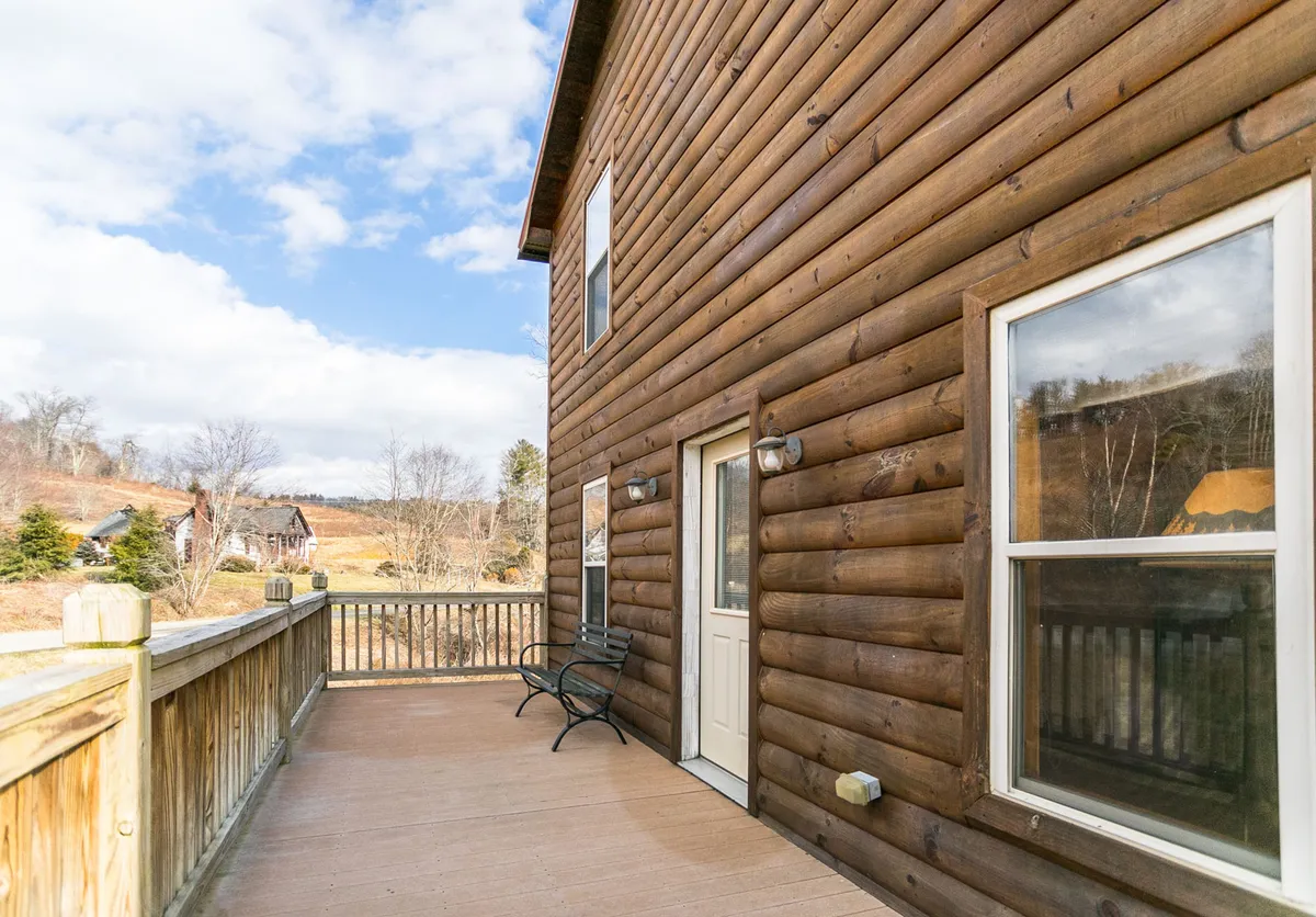 A peaceful exterior shot showing the wooden log cabin's deck with a sturdy railing and a bench, overlooking natural surroundings. This space invites guests to relax, enjoy morning coffee, or have quiet sunset chats while immersed in mountain scenery.