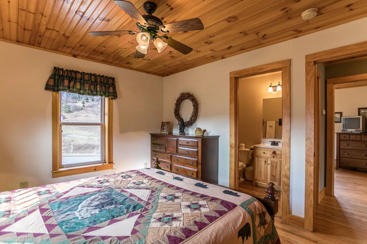 A warmly lit bedroom featuring a quilted bedspread, wooden dresser, and a window framing the scenic outdoors. The mood is inviting and tranquil, perfect for quiet moments or unwinding with a book while taking in the natural surroundings.