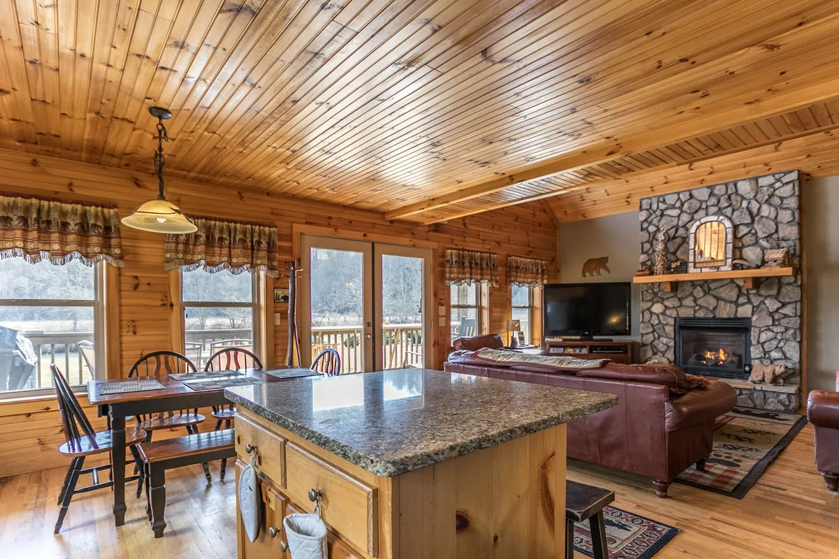 This image features the open-plan great room with a stunning stone fireplace as a focal point, plush leather sofas for gathering, a wooden dining table perfect for shared meals or games, and expansive windows flooding the space with light and offering spectacular views of the mountains and riverside.