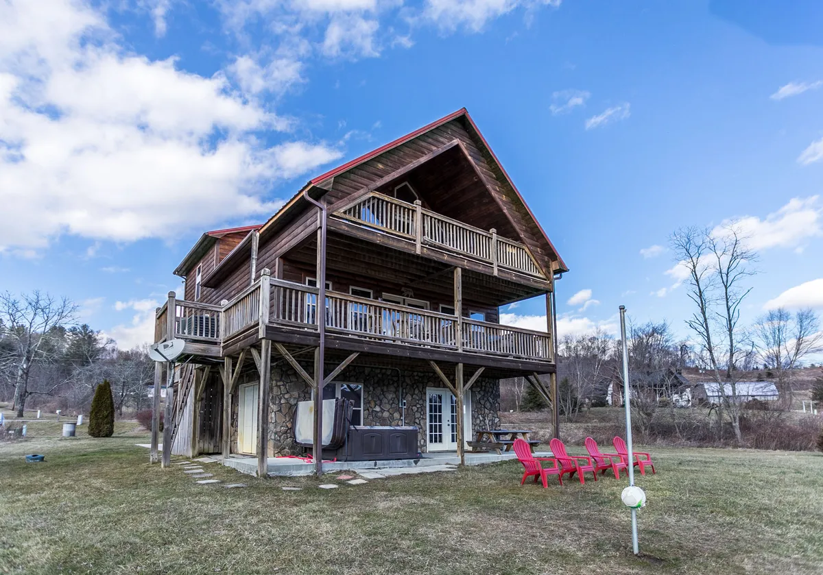 This exterior image captures the house structure with inviting red Adirondack chairs on the deck and a hot tub. The setting features a large deck area perfect for gatherings, surrounded by serene natural views, embodying both relaxation and the opportunity for outdoor activities.