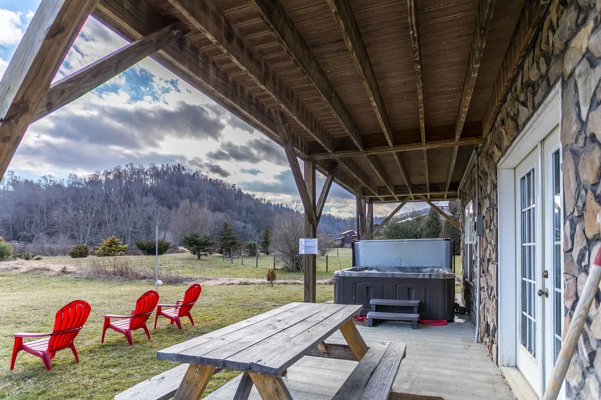 A rustic outdoor setting with a picnic table, red Adirondack chairs arranged around a stone fire pit, and a hot tub in a natural landscape. The scene is perfect for evening gatherings, storytelling, and enjoying the starry sky.