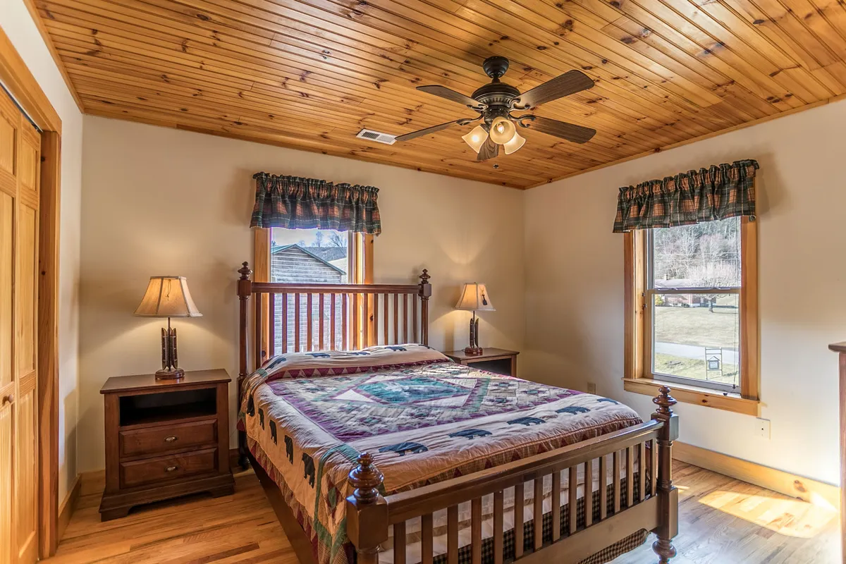 A warmly lit bedroom featuring a wooden bed frame, a decorative quilt, matching nightstands with lamps, a wooden ceiling, and windows with curtains allowing natural light to gently brighten the room.