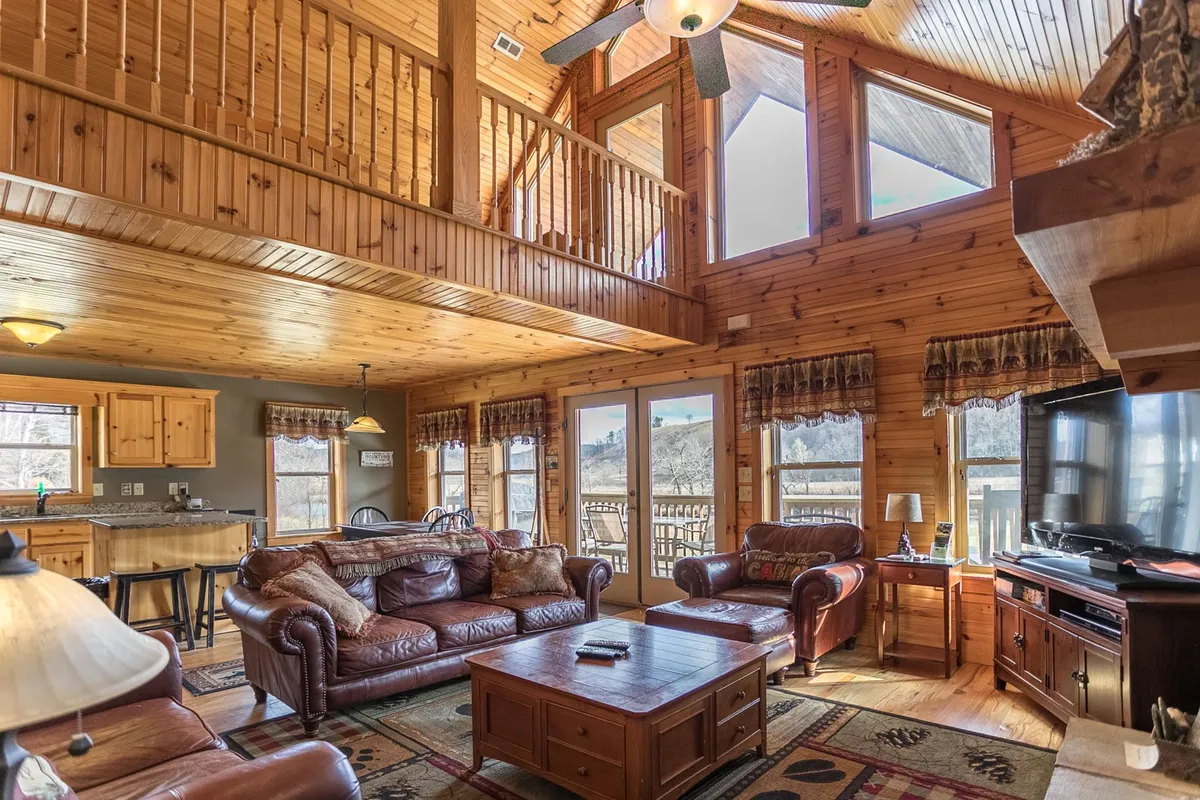 A different angle on the great room, highlighting the plush leather sofas arranged for conversation, a wooden coffee table ideal for games, and large windows showcasing sunset views. The wooden ceiling adds warmth and grandeur.