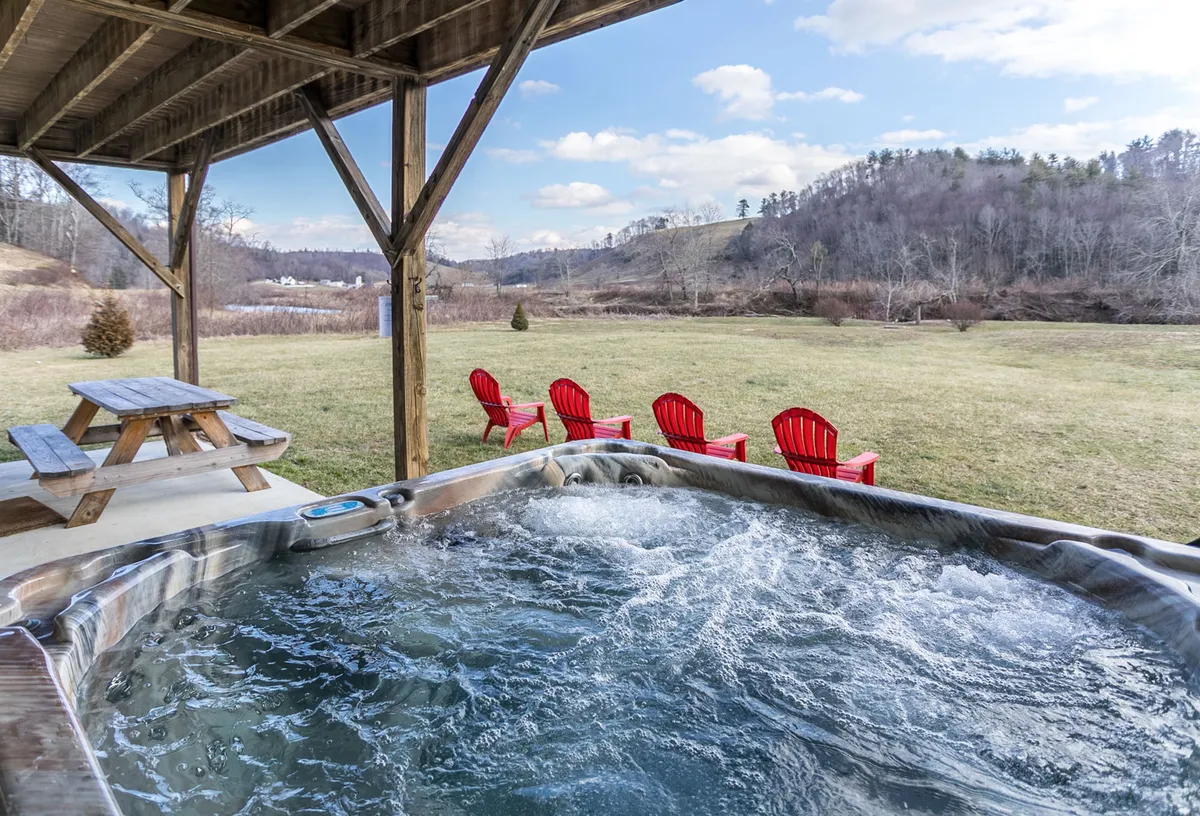 An exterior shot featuring a hot tub, red Adirondack chairs, a picnic table, and a scenic landscape in the background. The setup encourages relaxation, outdoor meals, and gathering with friends while enjoying the river and mountain setting.