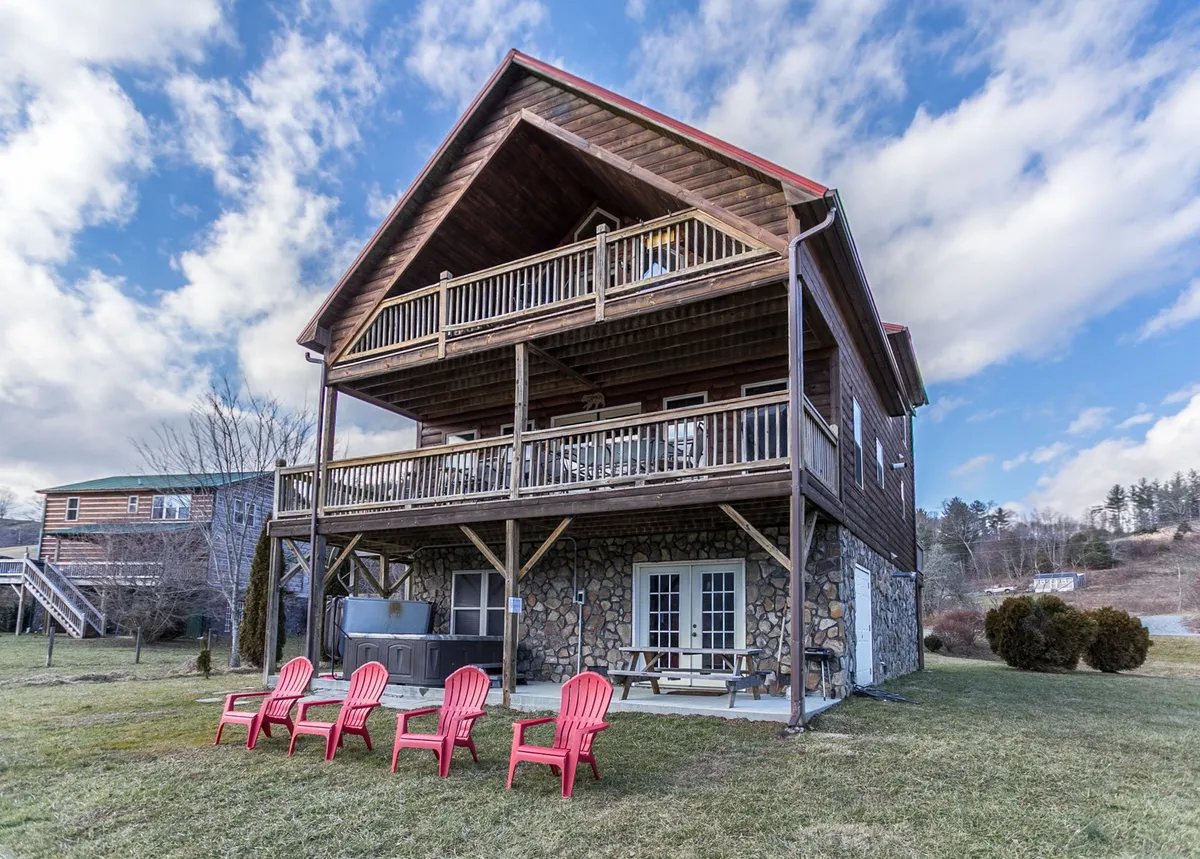 The image showcases the exterior of a charming wooden cabin with a spacious deck adorned with inviting red chairs, set against a backdrop of lush green grass and a rustic stone wall. The scene radiates relaxation, beckoning guests to sit outside for morning coffee, sunset views, or cozy gatherings under the stars.