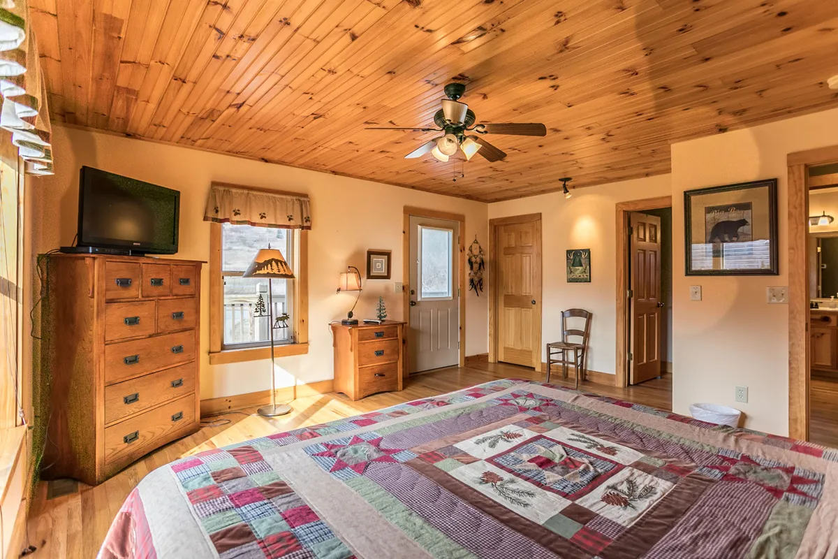 A cozy bedroom with a quilted bedspread, wooden furniture, a lamp beside the bed, and a television. Large windows invite the natural light and offer a relaxing view of the outdoors, hinting at a peaceful, private setting ideal for couples seeking a serene retreat.