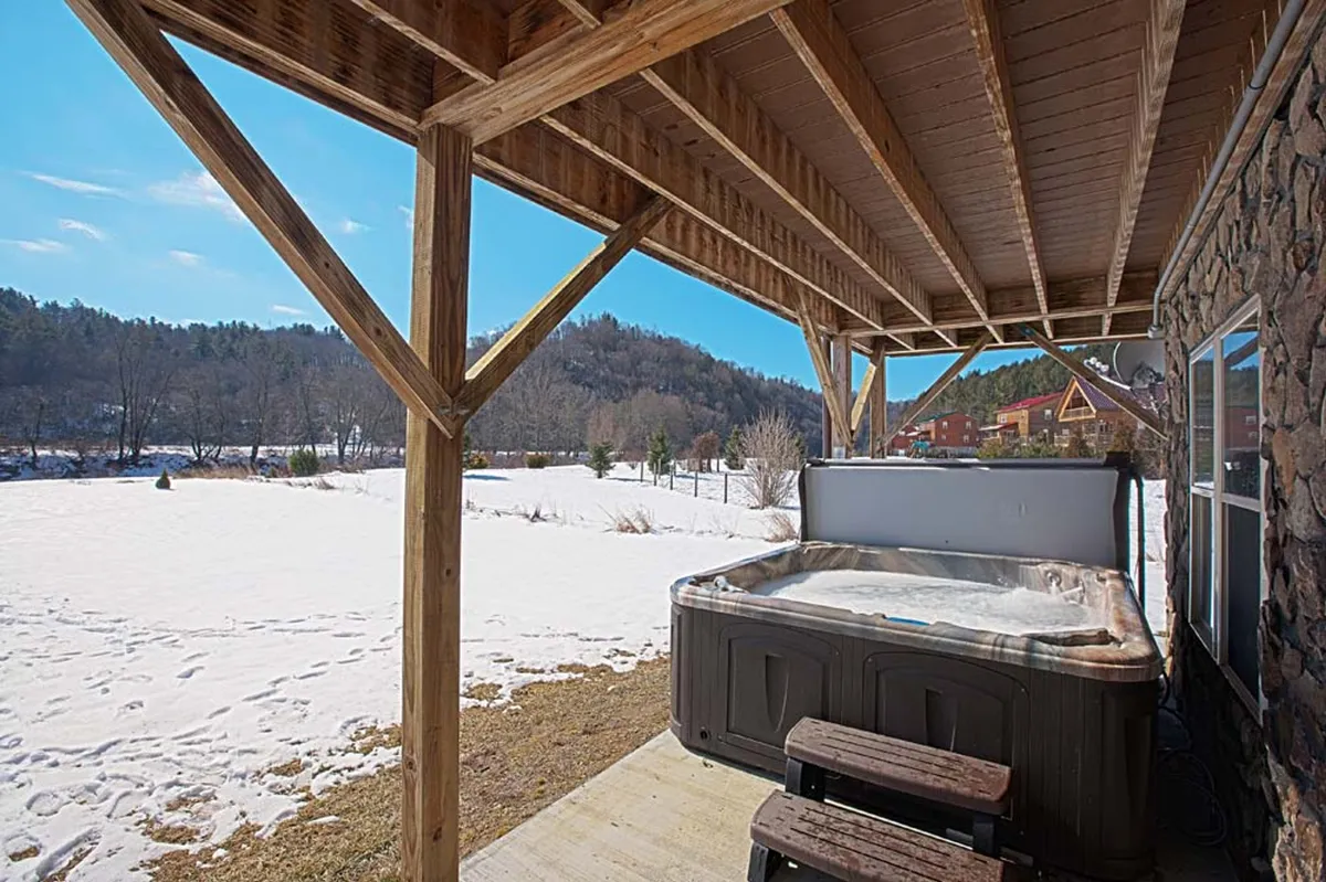 An inviting outdoor hot tub set beneath sturdy wooden beams, surrounded by a serene snow-covered landscape. The warm water steams against the chilly winter air, with the snowy backdrop visible beyond the deck railings.