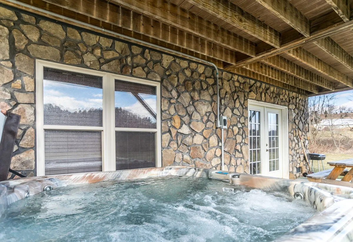 An inviting exterior view of a hot tub set against a stone wall, with double doors that hint at easy indoor-outdoor transitions. The tranquil mood and natural setting create an ideal spot for soaking and connecting under the stars.