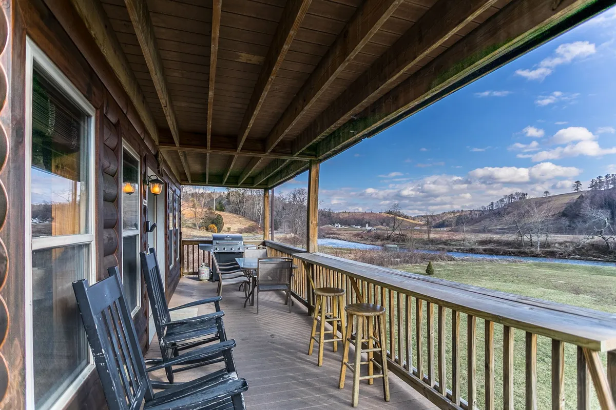 An inviting covered deck with wooden rocking chairs and bar stools overlooks the river and rolling hills. The grill is visible on the side, and the scene evokes the ideal setting for outdoor meals or morning coffee while enjoying scenic views.