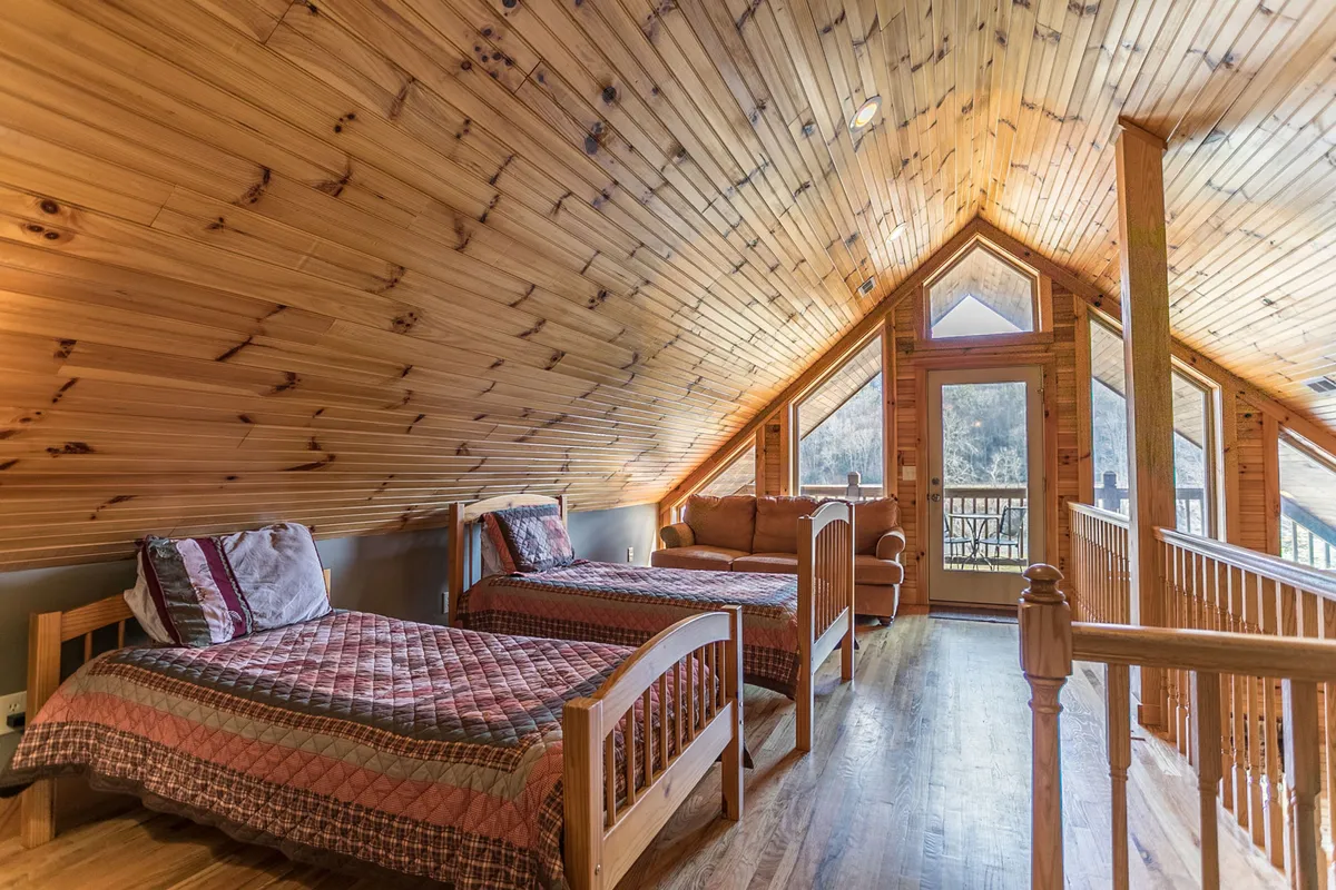 A spacious upper-level bedroom or loft area with wooden ceilings, large windows offering expansive mountain views, multiple beds, and a comfortable seating area for relaxing or sharing stories.